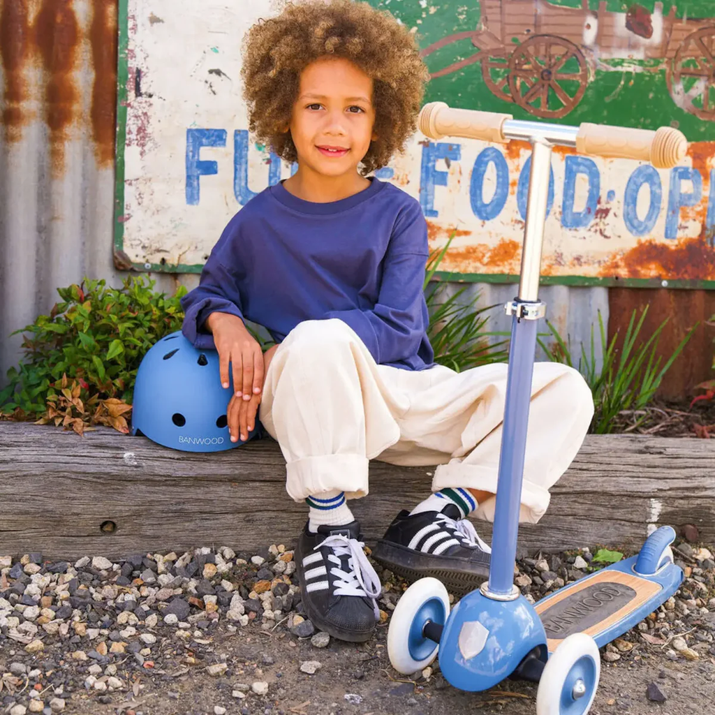 Young child with curly hair sits on a wooden bench, wearing a blue shirt and white pants. A blue helmet and scooter are beside them. Rustic sign in background.