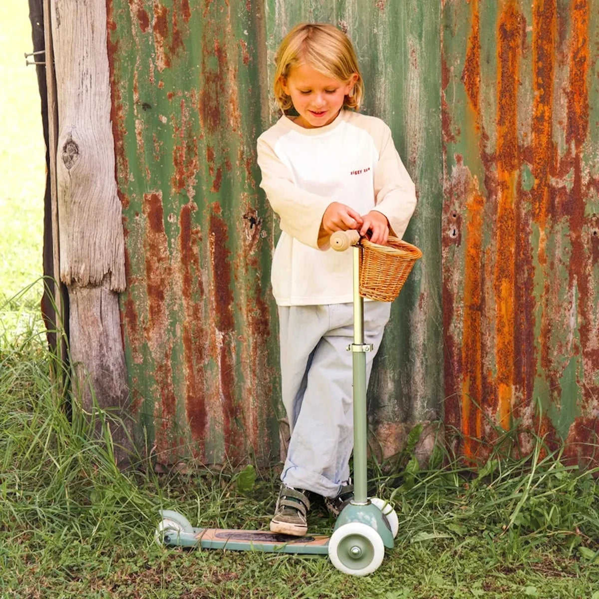 Young child standing with a sage green 3-wheeled scooter against a rustic, rusted metal wall outdoors.