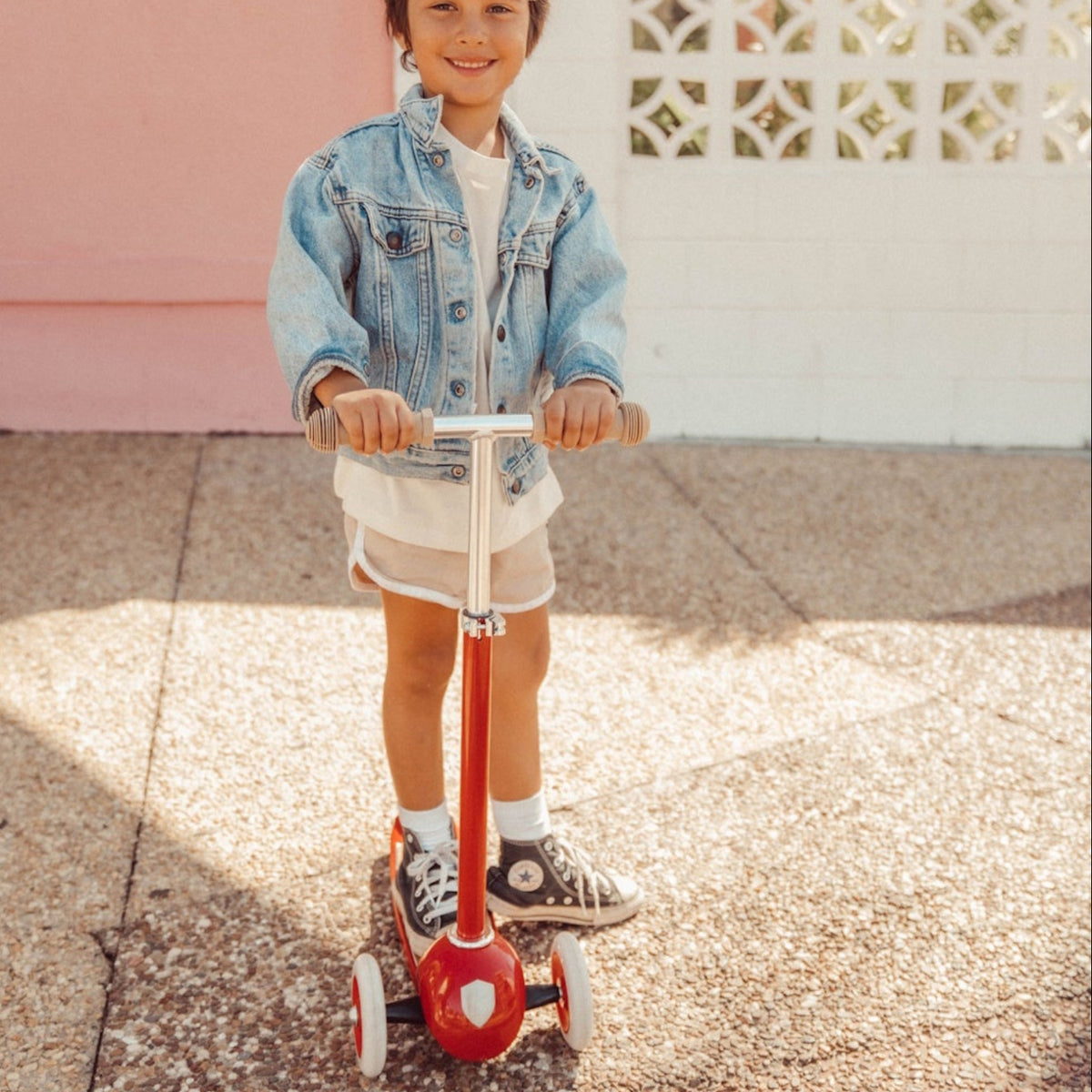 Smiling child in a denim jacket standing outdoors with a red three-wheeled kick scooter.