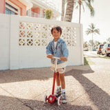 Young boy in denim jacket riding a red three-wheeled scooter on a sunny sidewalk.