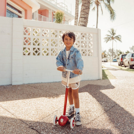 Young boy in denim jacket riding a red three-wheeled scooter on a sunny sidewalk.