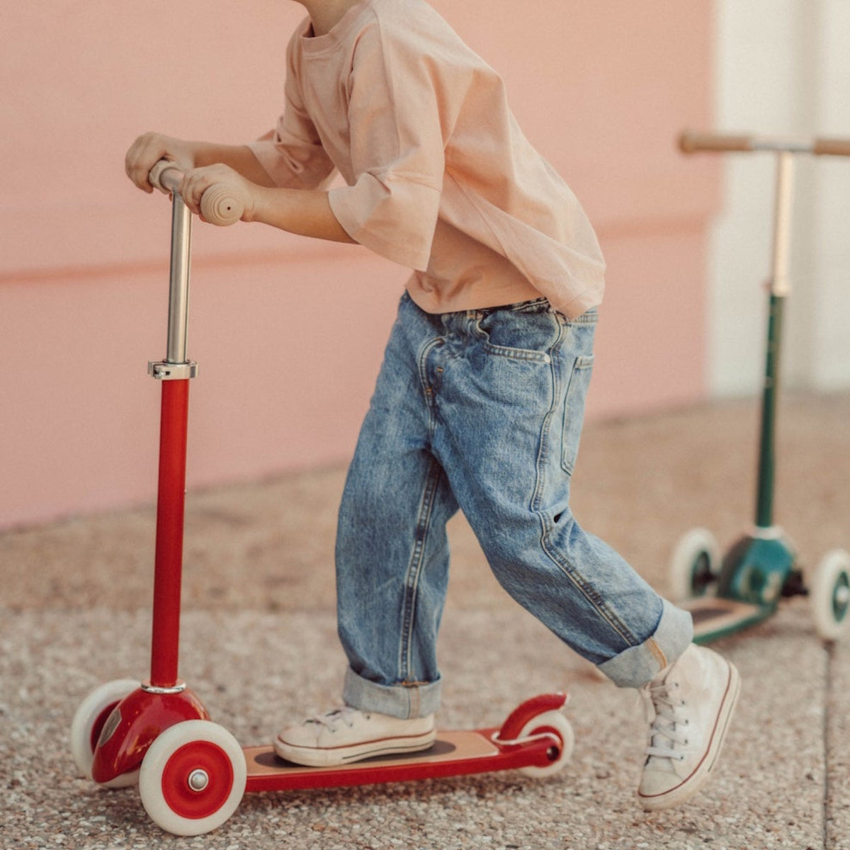 Child in cuffed jeans pushing a red three-wheeled kick scooter along a pavement.