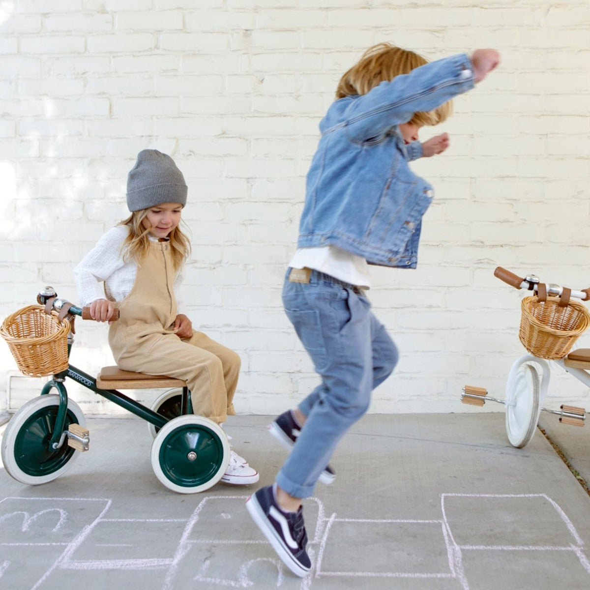 Two children playing outdoors; one sits on a green tricycle while another jumps near hopscotch grid.