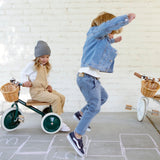 Two children playing outdoors; one sits on a green tricycle while another jumps near hopscotch grid.