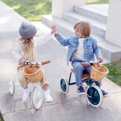 Two young children on Banwood tricycles giving each other a high five on a sidewalk outdoors.