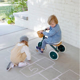Two young children playing on a sidewalk; one sits on a green tricycle while the other draws hopscotch in chalk.