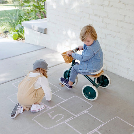 Two young children playing on a sidewalk; one sits on a green tricycle while the other draws hopscotch in chalk.