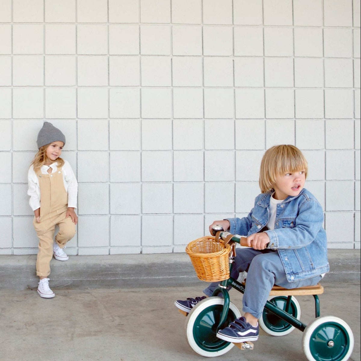 A young child in a denim jacket rides a green tricycle while another child leans against a tiled wall next to a white tricycle.