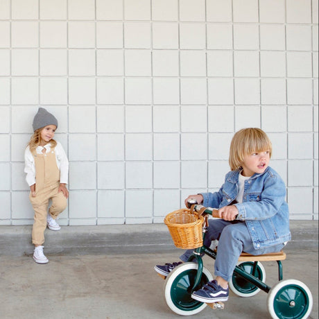 A young child in a denim jacket rides a green tricycle while another child leans against a tiled wall next to a white tricycle.