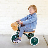 Young child in a denim jacket riding a green Banwood vintage tricycle against a white brick wall.