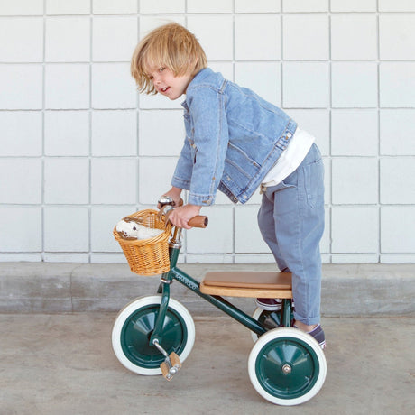 Child standing on a green Banwood tricycle with a plush toy in the wicker basket.
