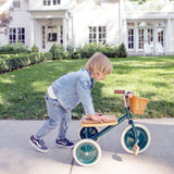 Child pushing a green Banwood tricycle along a sidewalk in front of a white house.