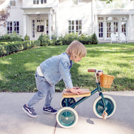Child pushing a green Banwood tricycle along a sidewalk in front of a white house.