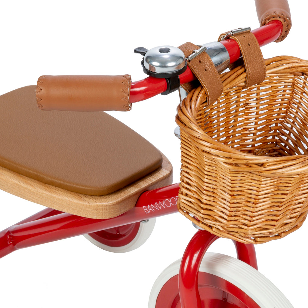 A classic children’s tricycle with a vibrant red steel frame, wooden pedals, removable push bar, and a wicker basket for timeless style and fun rides.