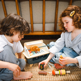 Two children sitting on the floor playing together with a wooden tabletop barbecue set.