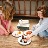 Two girls playing with a wooden barbecue set and serving play food on plates.