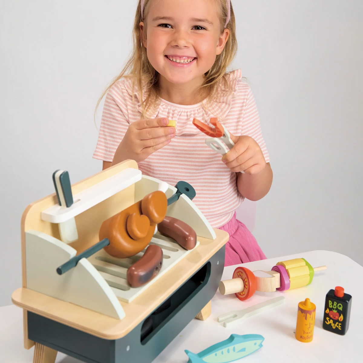 Smiling girl using wooden tongs to handle play food over a toy barbecue set.