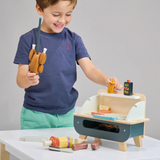 Young boy holding a toy rotisserie chicken skewer over a wooden barbecue play set.