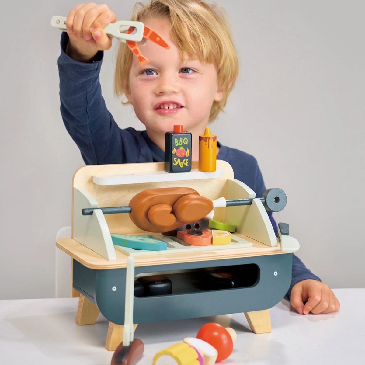 Young boy lifting a toy wooden prawn using tongs at a play barbecue grill set.