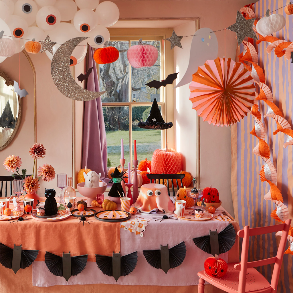 Festive Halloween table setup in pastel colors with pumpkins, paper bats, witch hats, and ghost decorations. A whimsical, playful atmosphere.