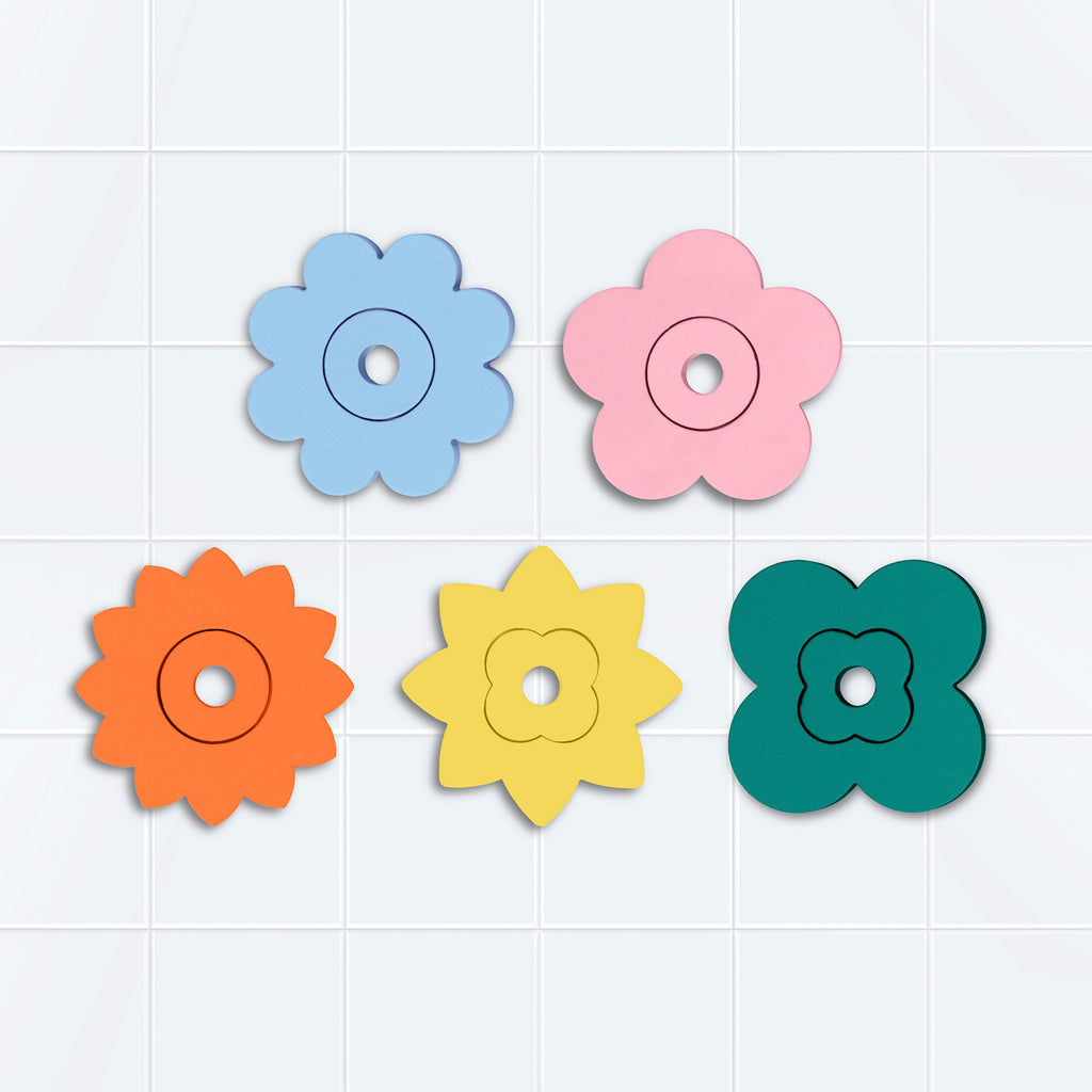 Five colorful flower-shaped coasters, in blue, pink, orange, yellow, and green, are arranged on a white tiled surface. The design is playful and vibrant.