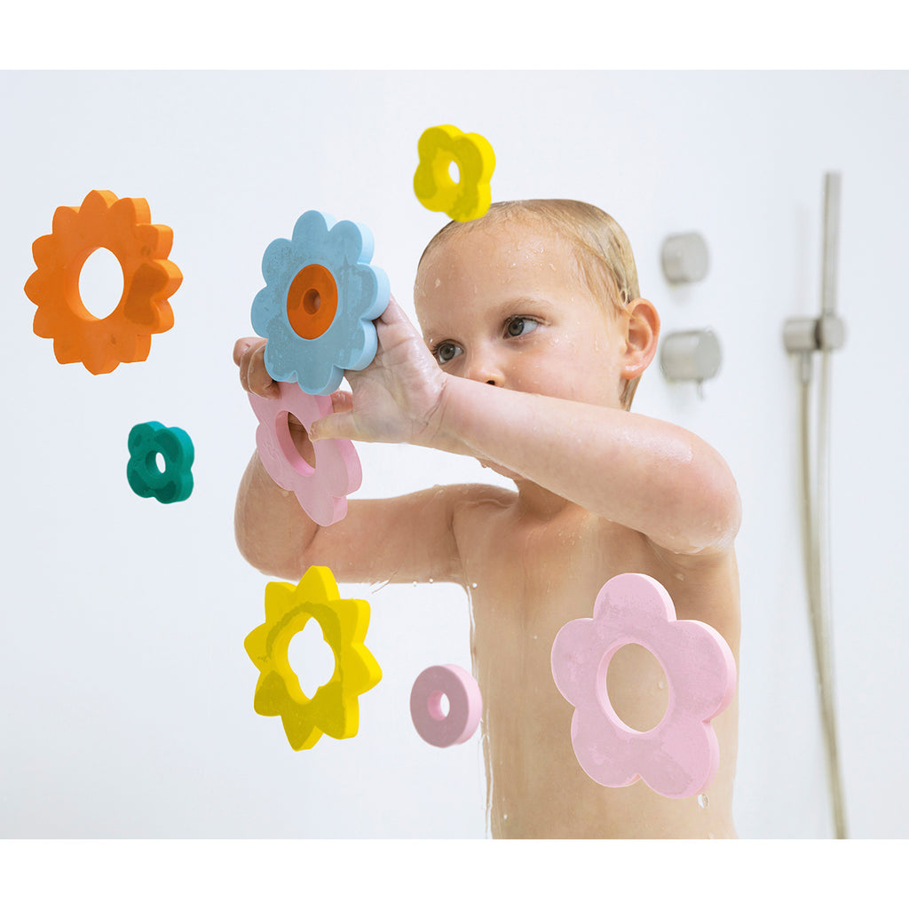 A baby in a bath, smiling and playing with colorful flower-shaped foam toys stuck to a glass surface. The mood is playful and joyful.