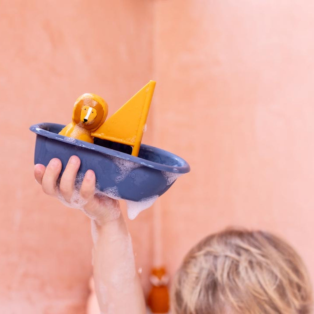 A child holds up a blue toy boat with a yellow sail and a small yellow figure. Soap suds cover the boat, set against a peach-colored background. Playful tone.