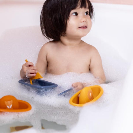 A toddler looks away while playing with toy boats in a bubble bath.
