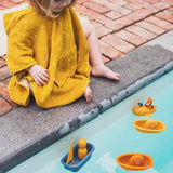 A toddler in a yellow hooded poncho sits by a pool watching toy boats float in the water.