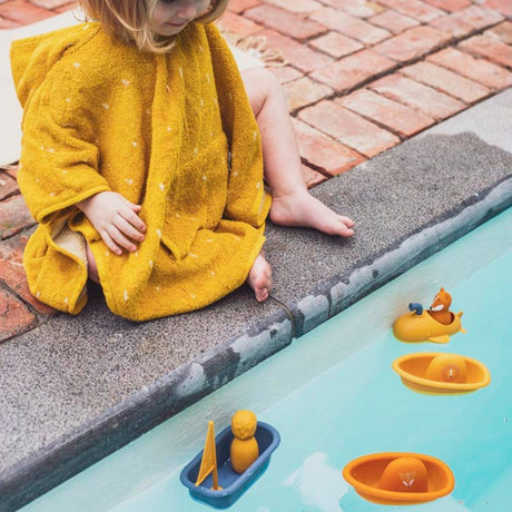 A toddler in a yellow hooded poncho sits by a pool watching toy boats float in the water.