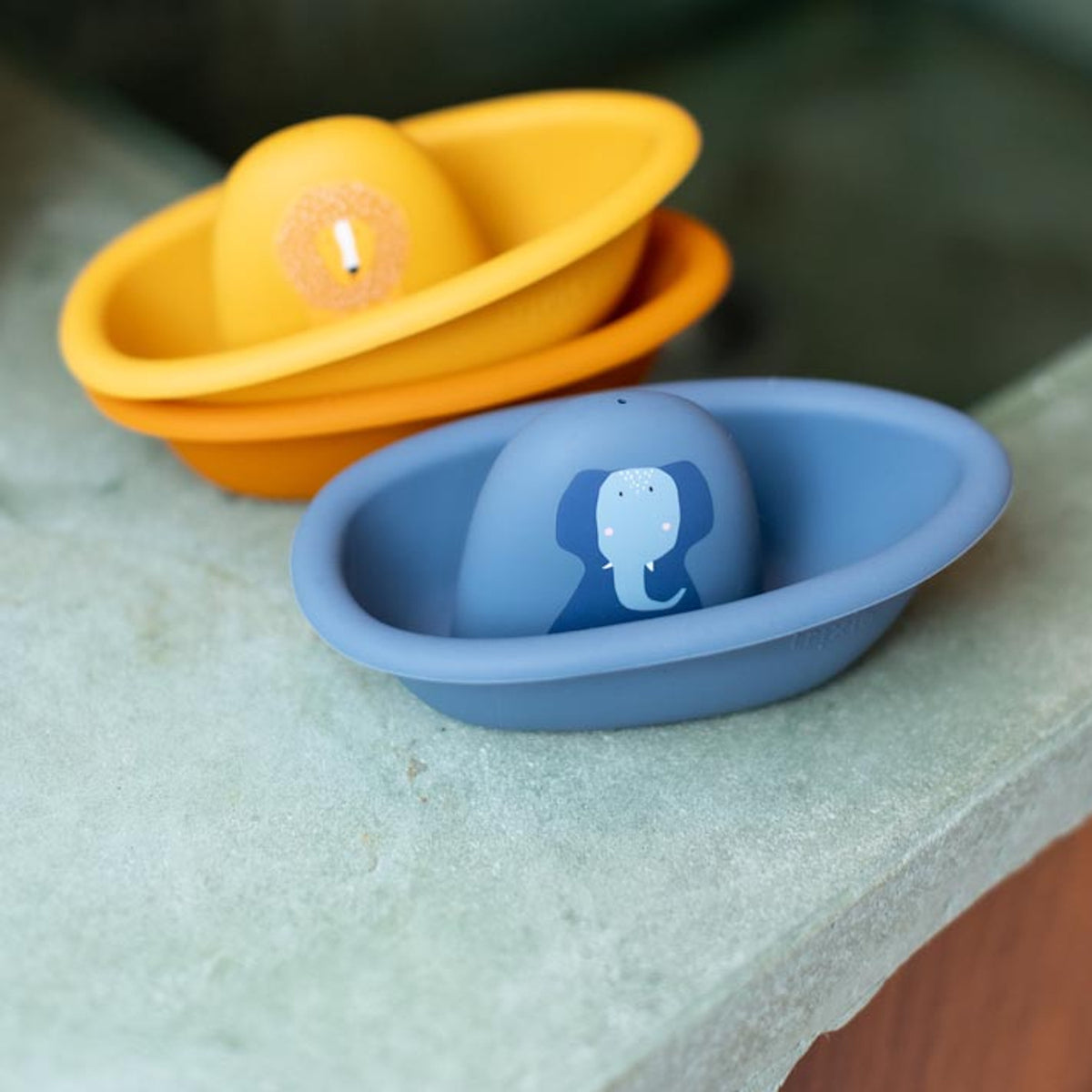 A blue toy boat with an elephant print next to stacked yellow and orange toy boats on a ledge.