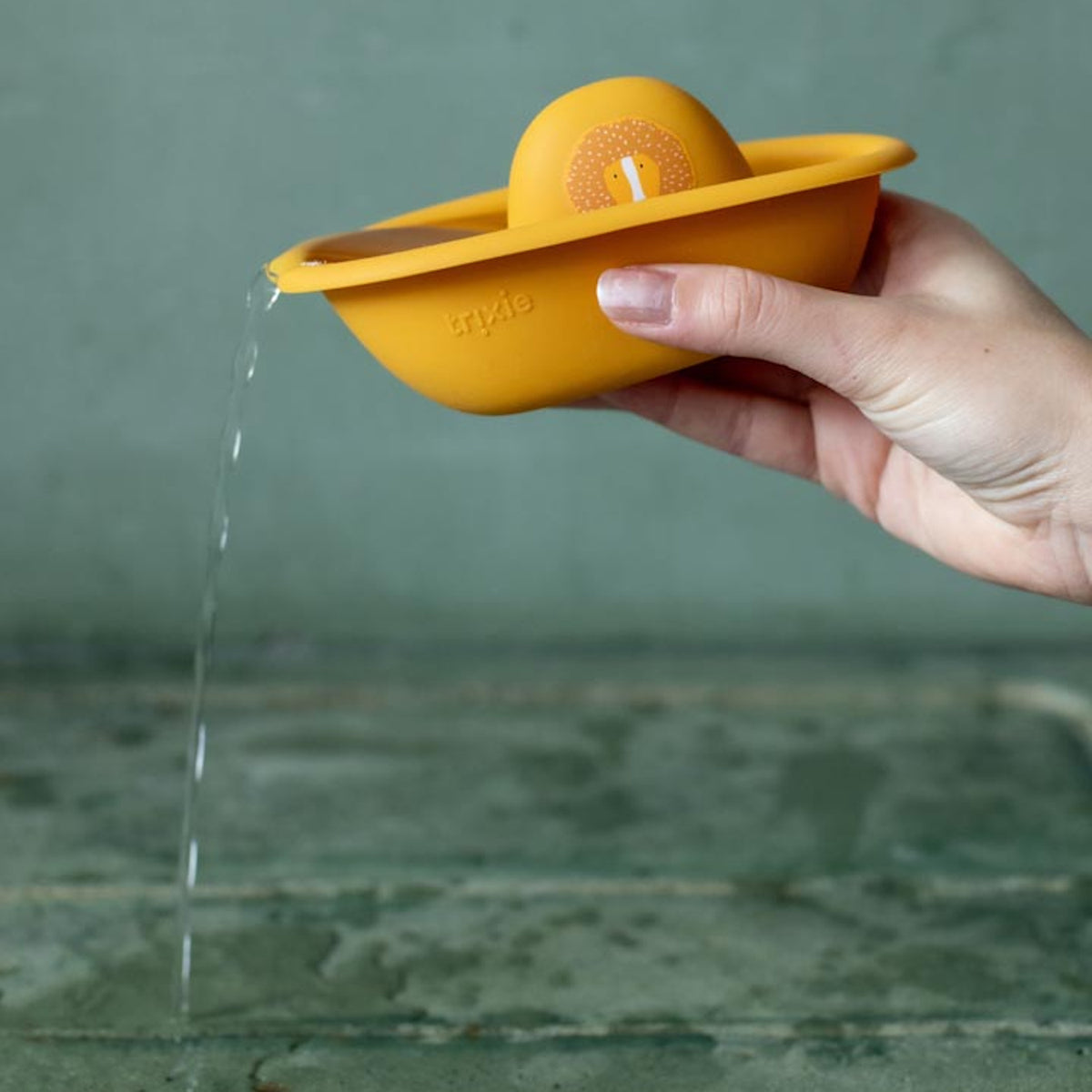 A hand pouring water out from a yellow toy boat with a lion face illustration.