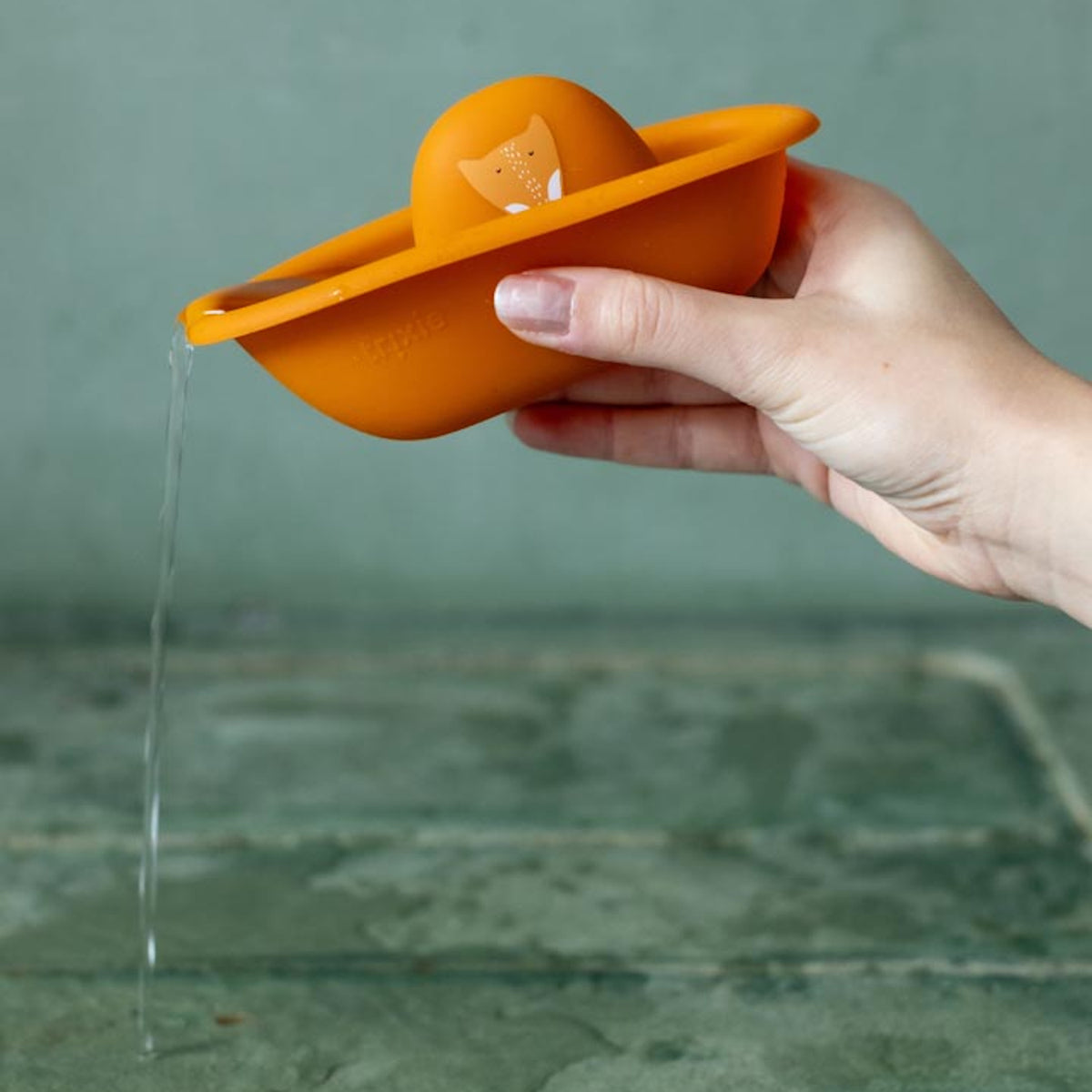 A hand holding an orange silicone fox bath toy boat as water pours out of its spout.