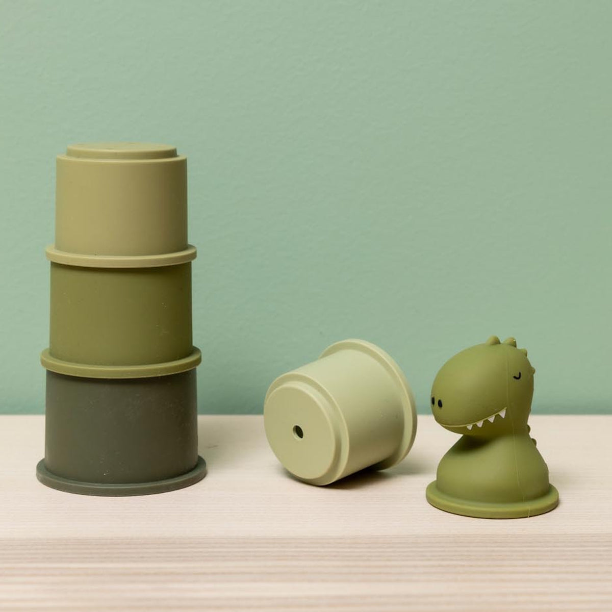 Green dinosaur bath toy with matching sage and olive green stacking cups for toddlers.