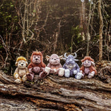 Five plush monster and creature toys lined up sitting in a row on a large fallen tree trunk in a forest.