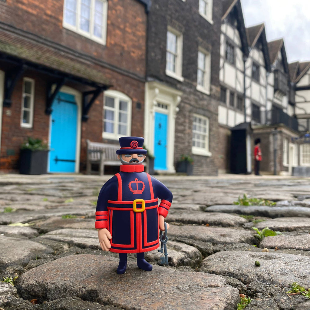 A toy figure of a Beefeater in blue and red stands on cobblestones with historic buildings and a vibrant blue door in the background. The scene feels whimsical.
