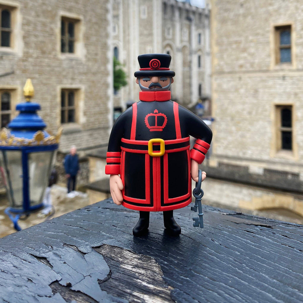 Toy figure of a Beefeater, in black and red uniform, holding keys, stands on a stone ledge with the Tower of London's historic walls in the background.