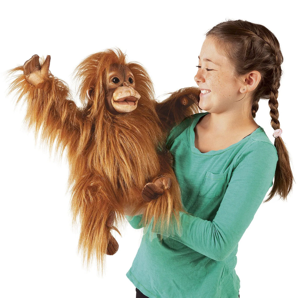 A smiling girl in a green shirt holds a realistic orangutan puppet. The puppet has long, orange fur and an expressive face, creating a joyful scene.