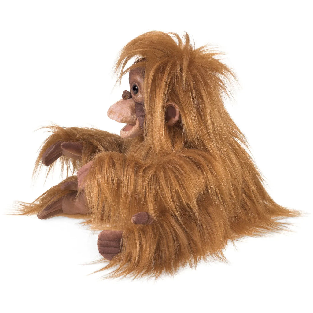 A plush orangutan toy with long, shaggy orange fur sits facing left. Its detailed facial features and relaxed pose convey a playful, whimsical tone.