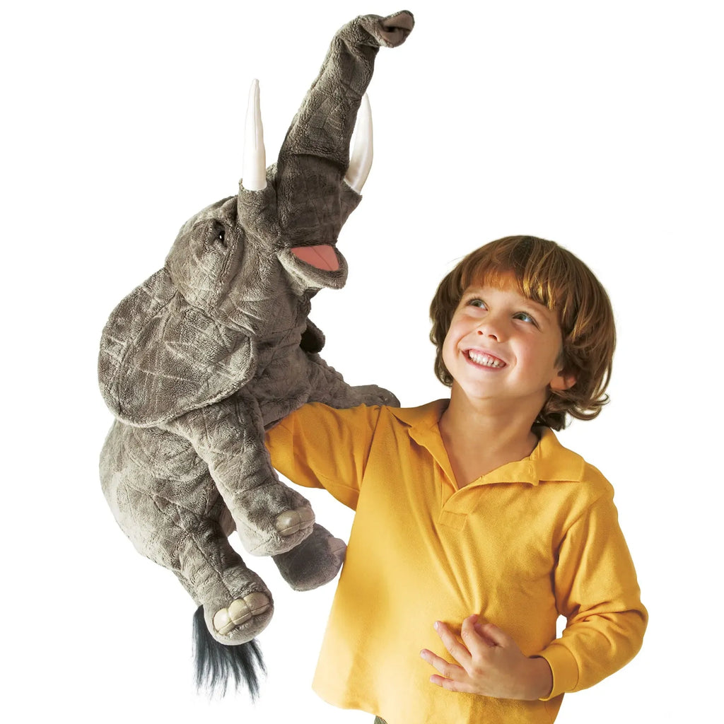A smiling young child in a yellow shirt holds a large plush elephant puppet. The elephant's trunk is raised, creating a joyful and playful atmosphere.