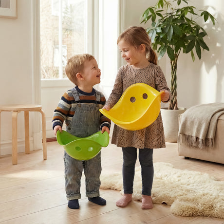 Two young children standing in a bright room holding a green and a yellow Bilibo toy, smiling at each other.