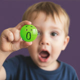 A young boy mimics the surprised expression of a green Bilibo Game Node toy he is holding against a purple background.