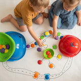Two young children playing a tabletop game with colorful Bilibo Game Shells and chips on a hand-drawn paper game board.