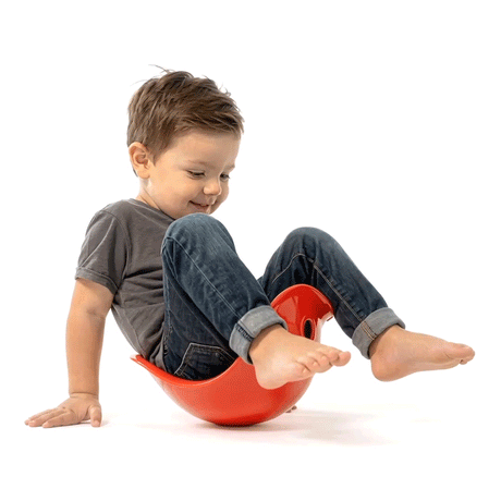 A smiling toddler in a grey shirt and jeans balancing barefoot on a red Bilibo toy on white background.