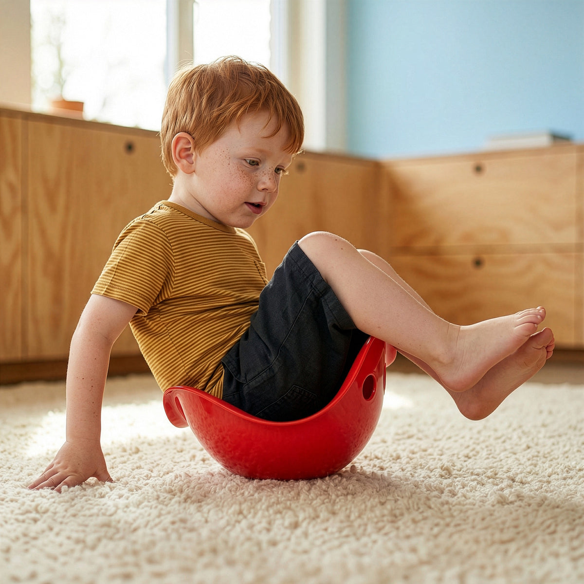 A young boy with red hair sits and balances in a red Bilibo seat on a plush rug in a sunlit playroom.