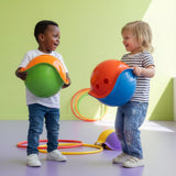 Two smiling toddlers holding colorful Bilibo MIDI sensory toys in a playroom with hula hoops on a green background.