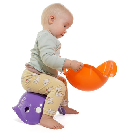 Toddler sitting on a purple Bilibo toy while holding an orange Bilibo, showcasing open-ended creative play for kids.