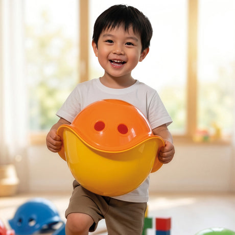 Smiling young boy holding an orange and yellow Bilibo toy, designed to look like a face, in a bright playroom.