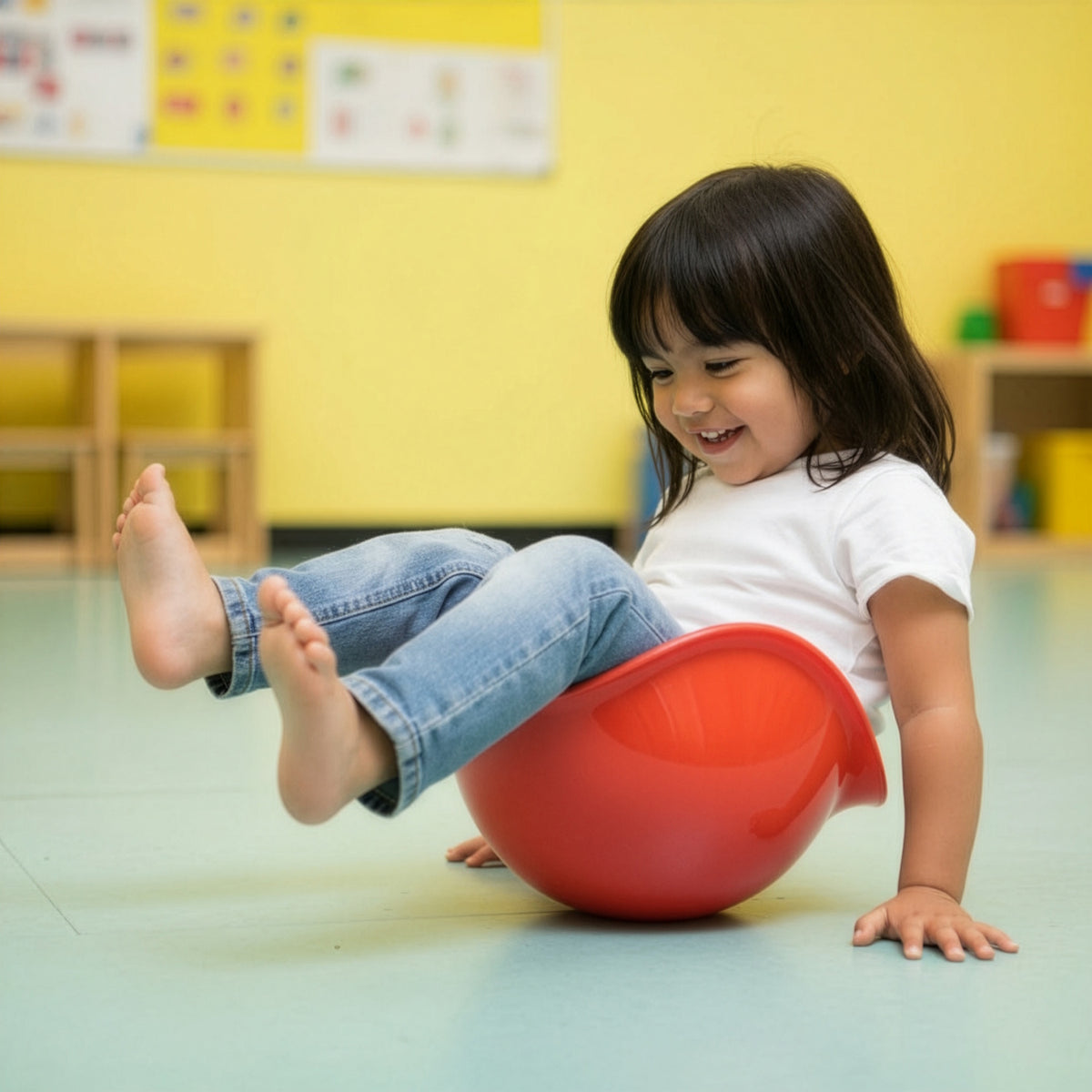 A smiling young girl playing and spinning in a red Bilibo seat on a classroom floor.