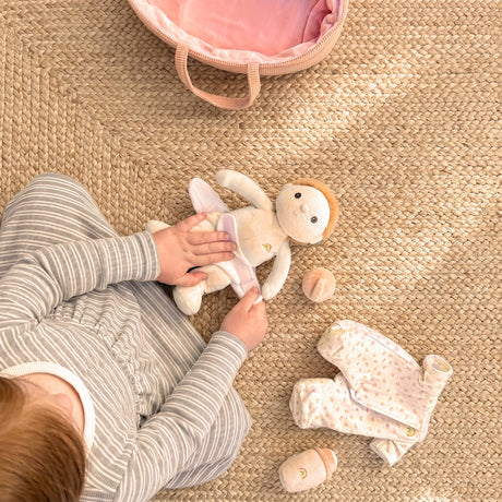 A child's hands changing a Dinky Dinkum doll on a woven rug next to doll clothes and a pink bag.